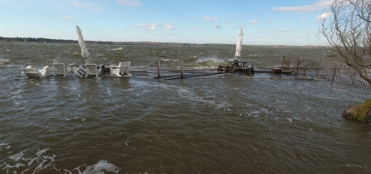 Coastal floodwater covering a patio: white chairs and a picnic table stand in moving brown water up to their seats, with a submerged dock, a half-buried fence, and the far shoreline behind them.
